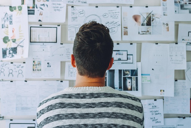 Man in striped sweater facing a wall covered with papers, sketches, and notes for brainstorming or planning