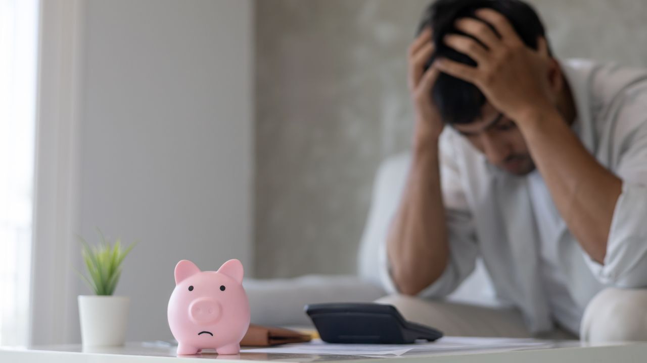 Stressed man holding his head while looking at bills on a table with a sad pink piggy bank in the foreground.