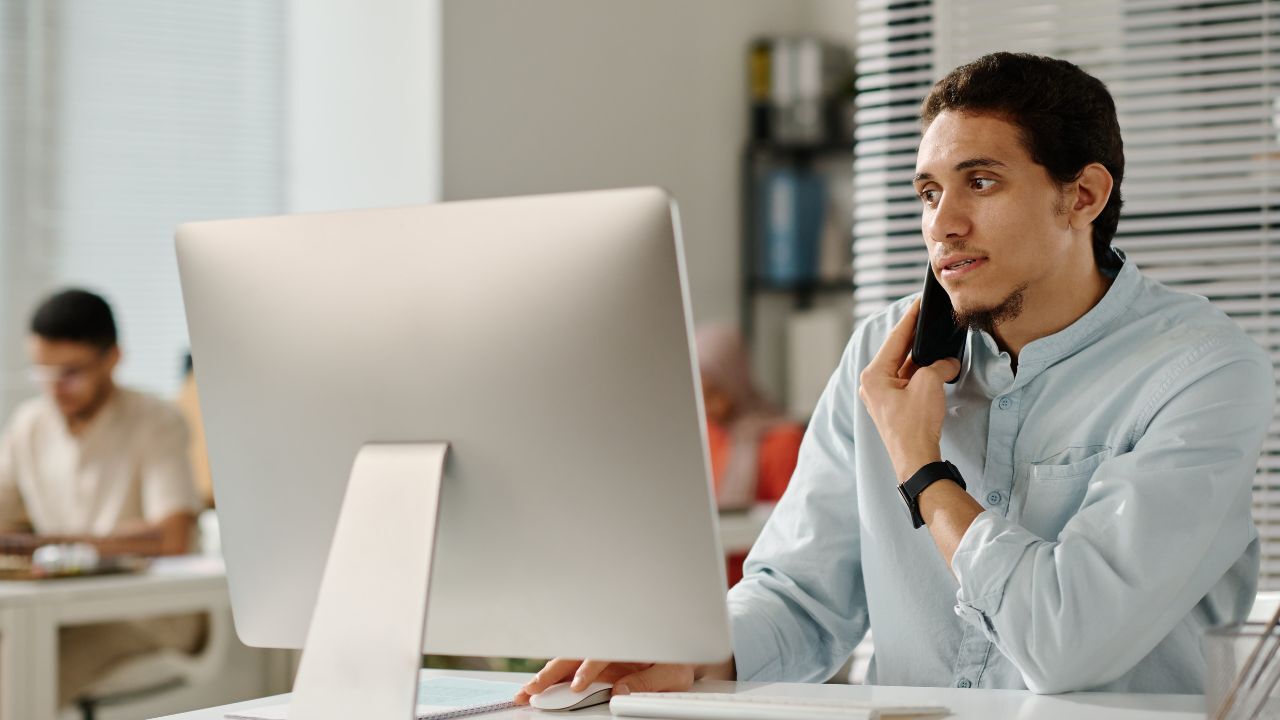 Young man sitting at a desk, talking on the phone while looking at a computer screen in an office.